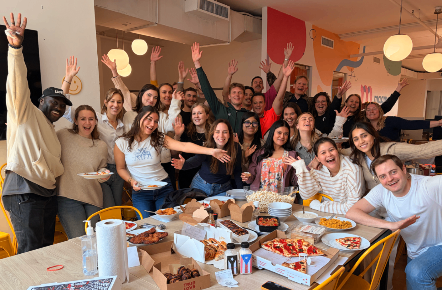 Office community pizza party with a large group of coworkers cheering behind a table full of food