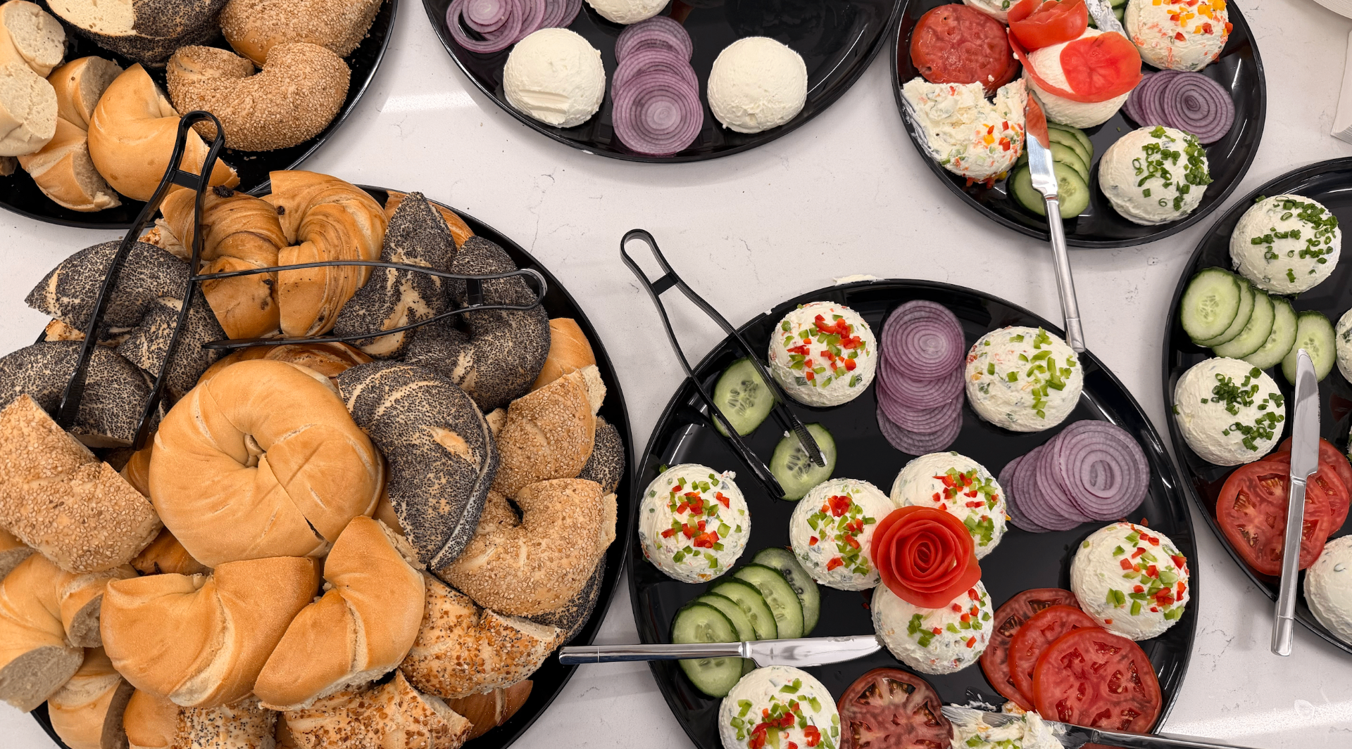Bagel and cream cheese catering spread arranged by Dixie, with bagels, spreads, tomatoes, cucumber and red onion on black plates